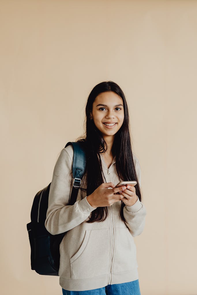 Teenage girl smiling while using smartphone, wearing a backpack, against a neutral background.